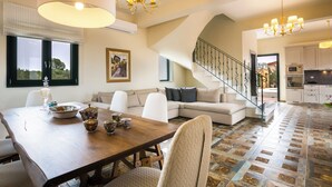 Un'ampia zona giorno con un tavolo da pranzo in legno, divani bianchi, una scala e una cucina sullo sfondo.