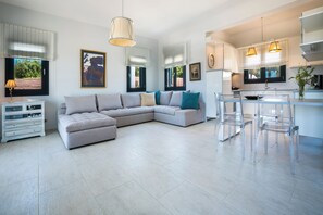 A modern living room with a large L-shaped sofa, a dining area with clear chairs, a kitchen with pendant lights, and a white cabinet.