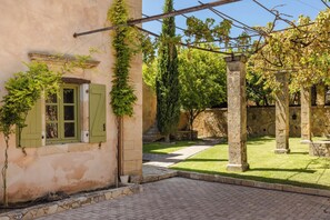 A courtyard with a stone wall, a pergola with climbing vines, and a grassy area.