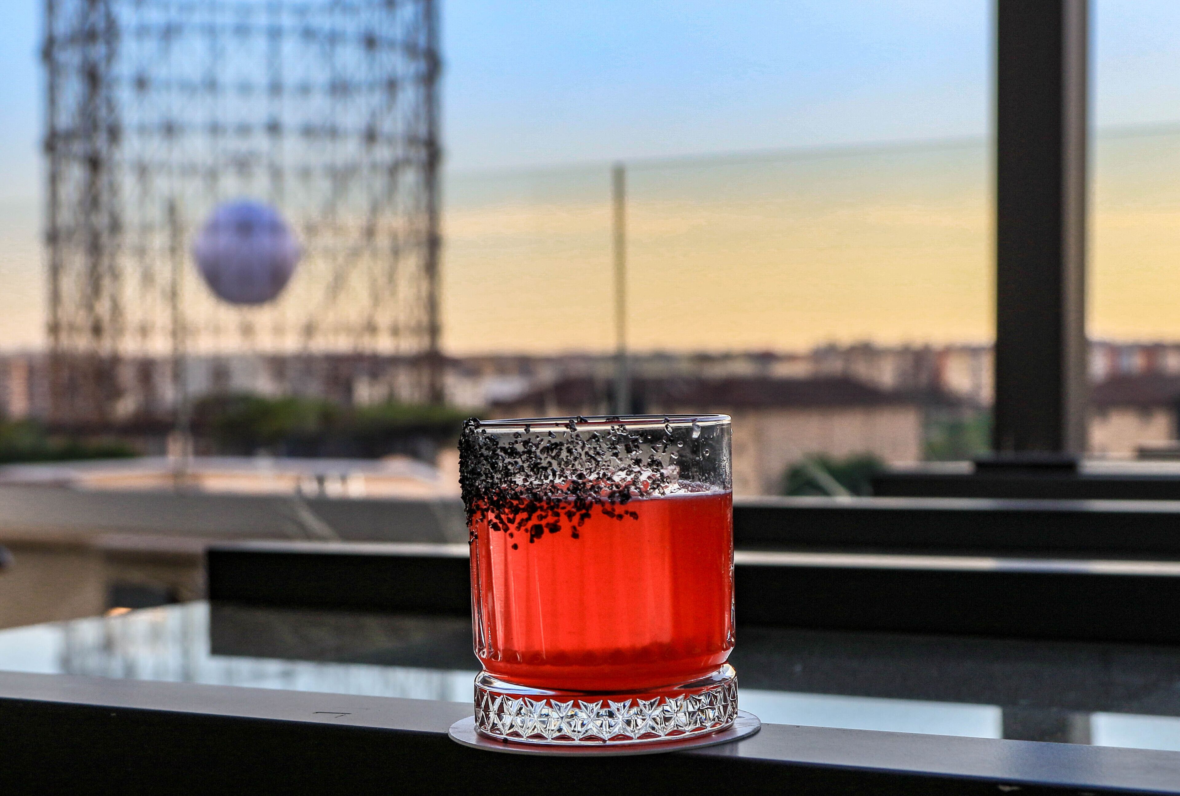 Et glas med rød væske på en balkon med udsigt til en bygning og en klar himmel.