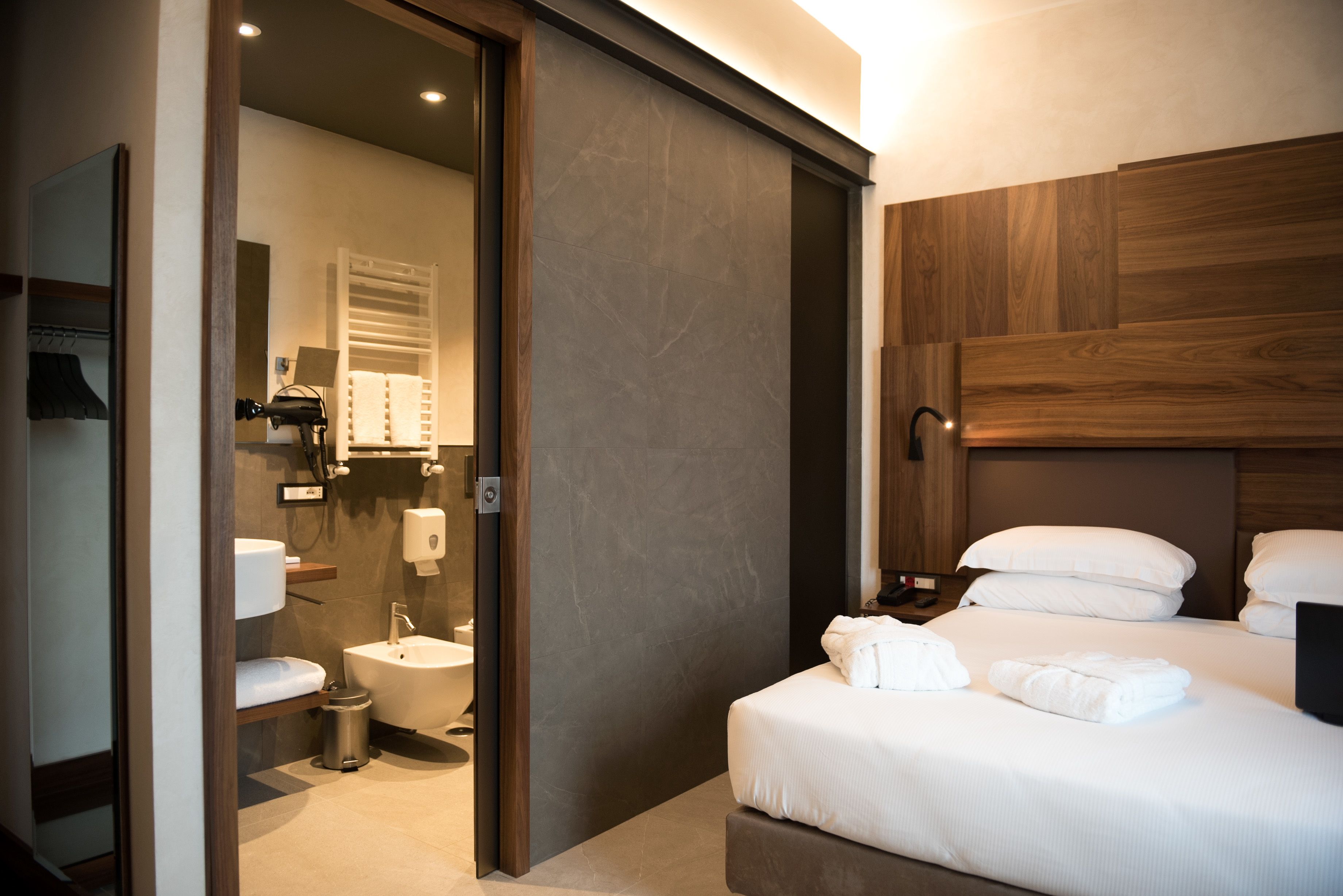 A modern hotel room with a bed, bedside table, and a bathroom with a toilet, sink, and towel rack.