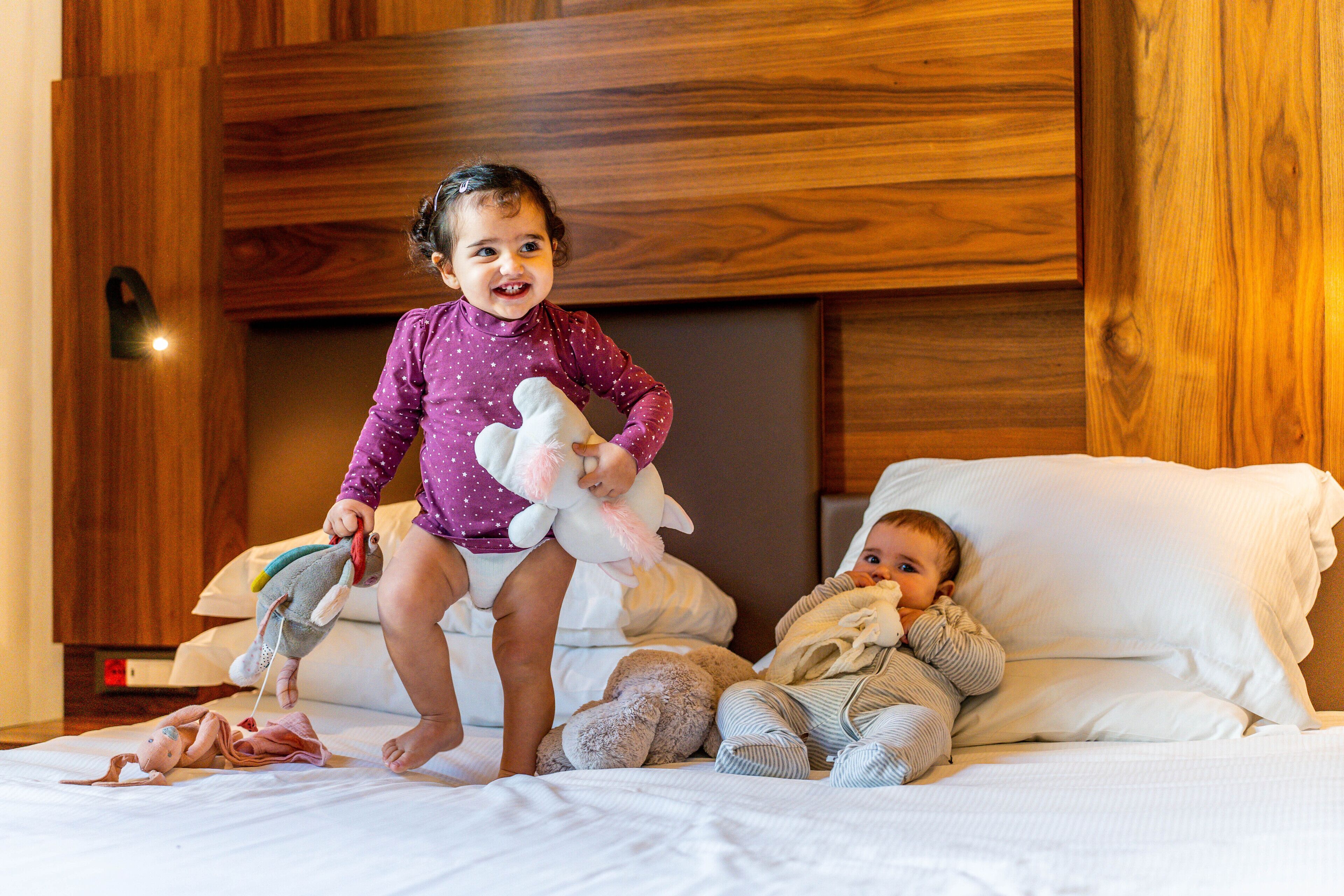 Niño jugando con un peluche en una cama con cabecera de madera y ropa de cama blanca.