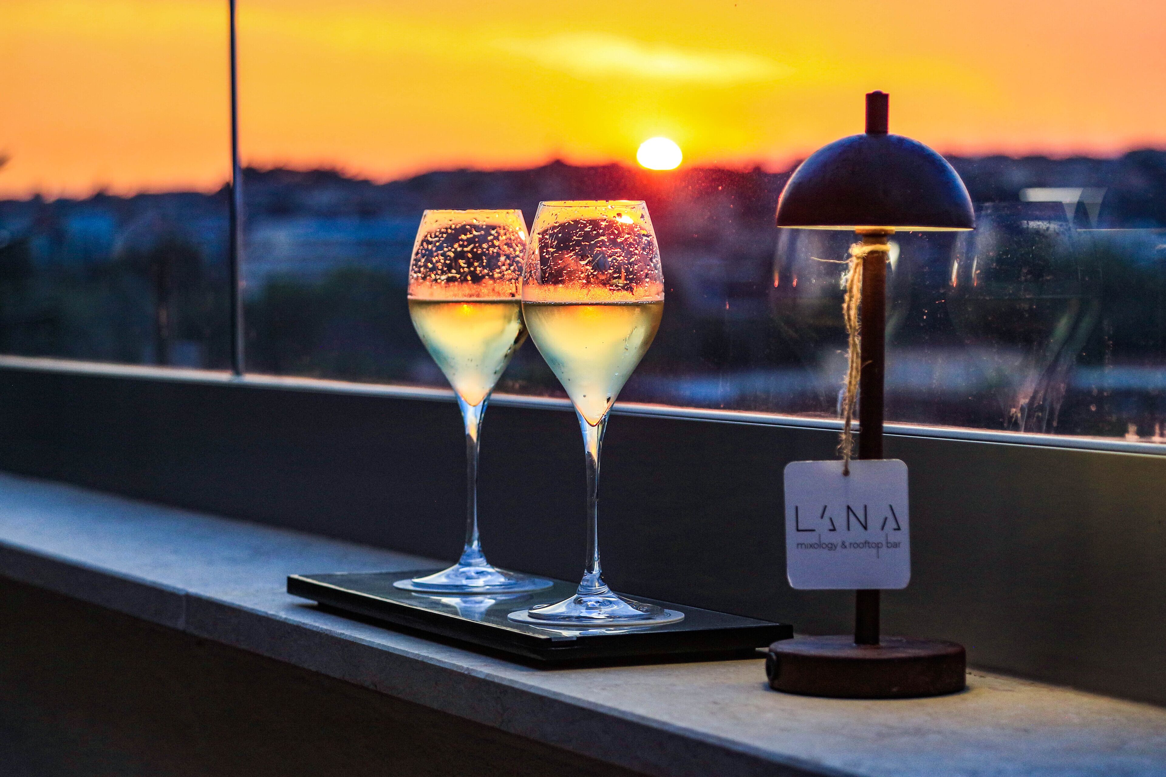 Deux verres contenant un liquide transparent, peut-être un cocktail, sont posés sur un plateau à côté d’un panneau indiquant « L’NA » et précisant : « musée et bar sur le toit ».