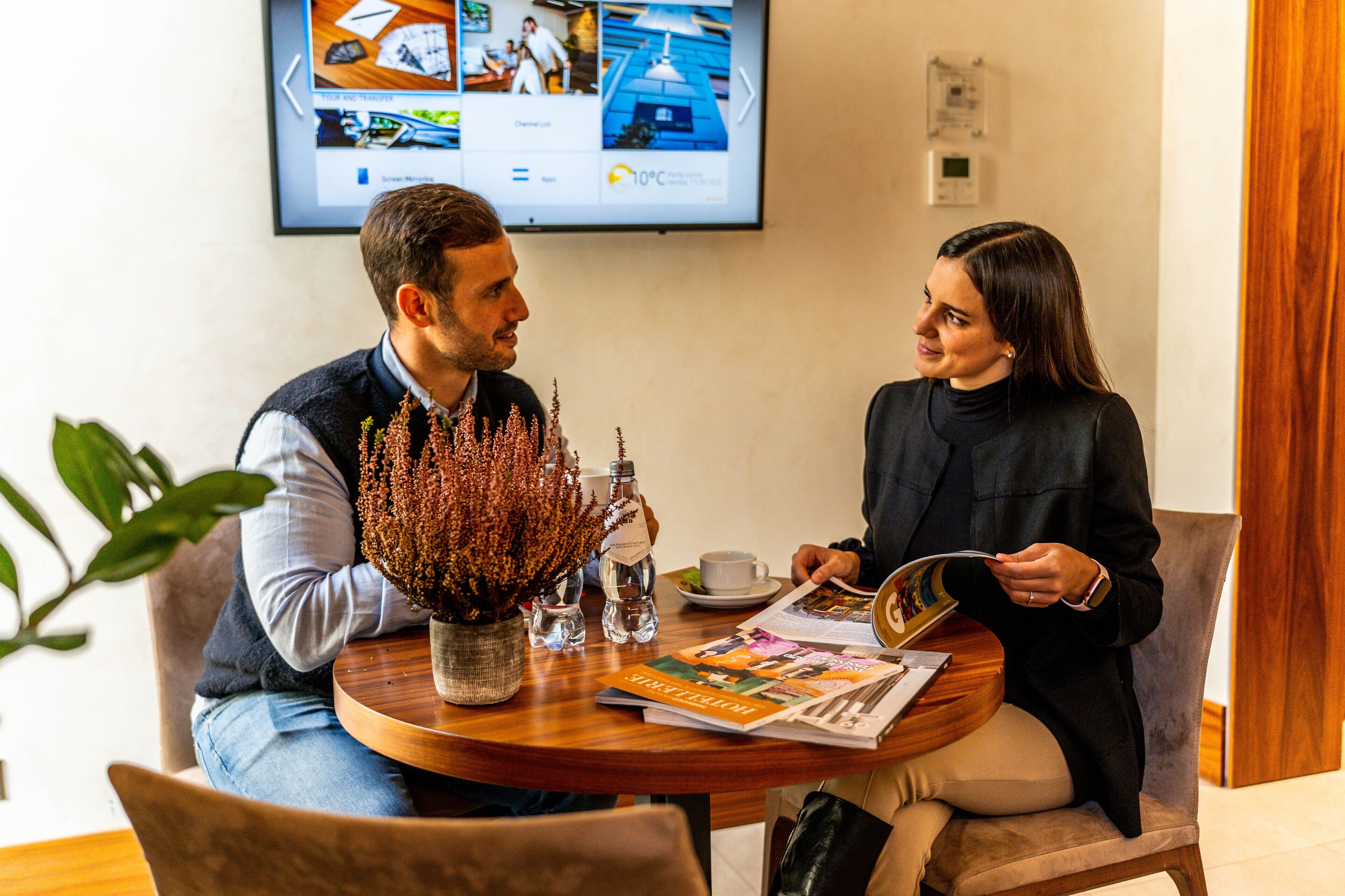 A man and a woman sitting at a round wooden table with a magazine, a small potted plant, and a cup of coffee.