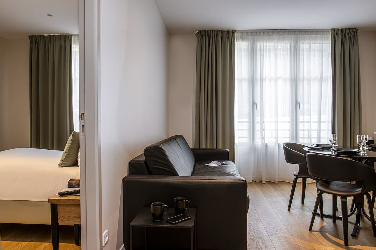 A hotel room with a bed, a sofa, a dining table with chairs, and a small side table with a remote control and a mug.