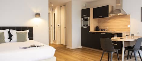 A modern hotel room with a bed, bedside table, and a small kitchenette.