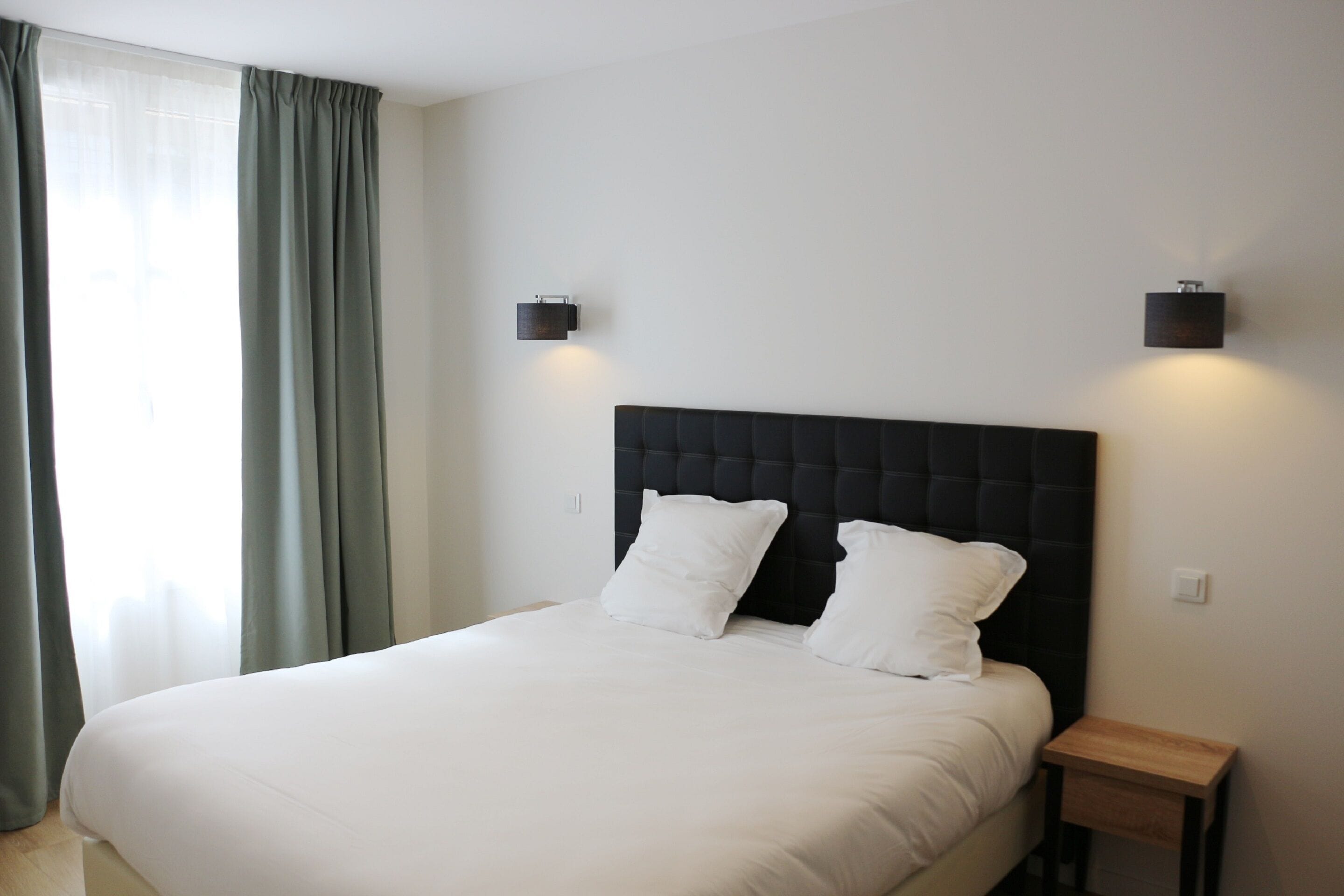 A neatly made bed with white linens and two pillows, a dark headboard, and two wall-mounted light fixtures.