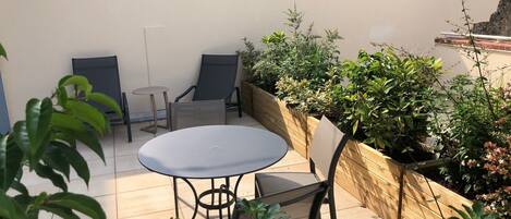Un espace patio avec une table ronde et deux chaises, entouré de plantes et d’un jardinière en bois.