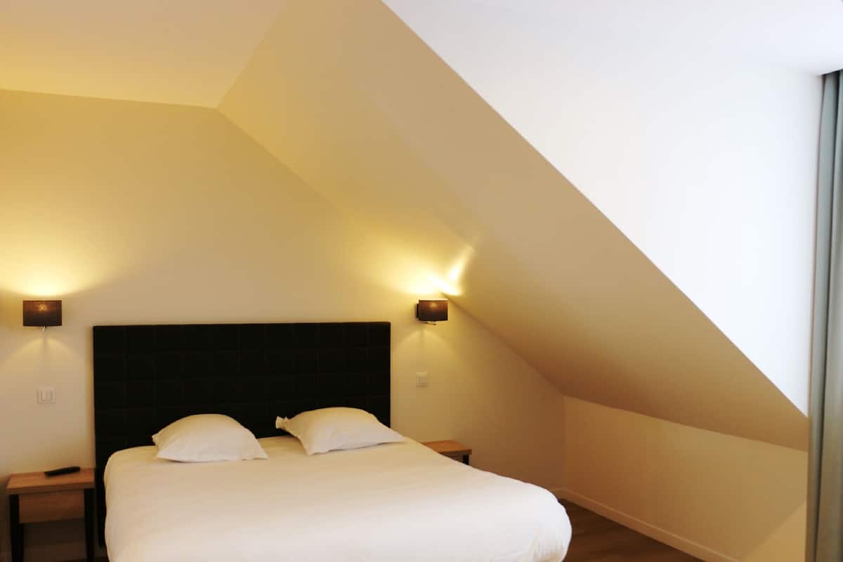 A bedroom with a high ceiling, a large bed with white linens, two bedside lamps, and a dark headboard.