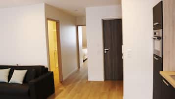 A modern apartment with a black sofa, wooden flooring, a kitchen with a built-in oven, and a door leading to another room.