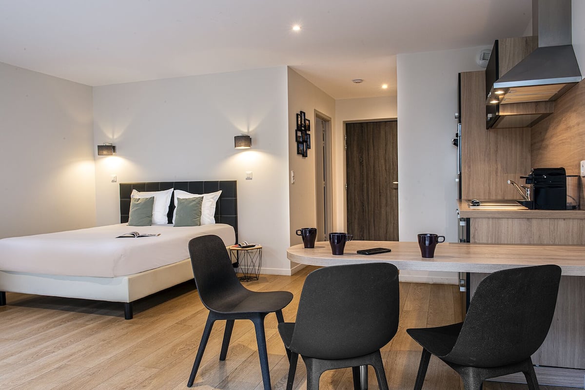 A modern hotel room with a bed, two chairs, a small table, and a kitchenette.