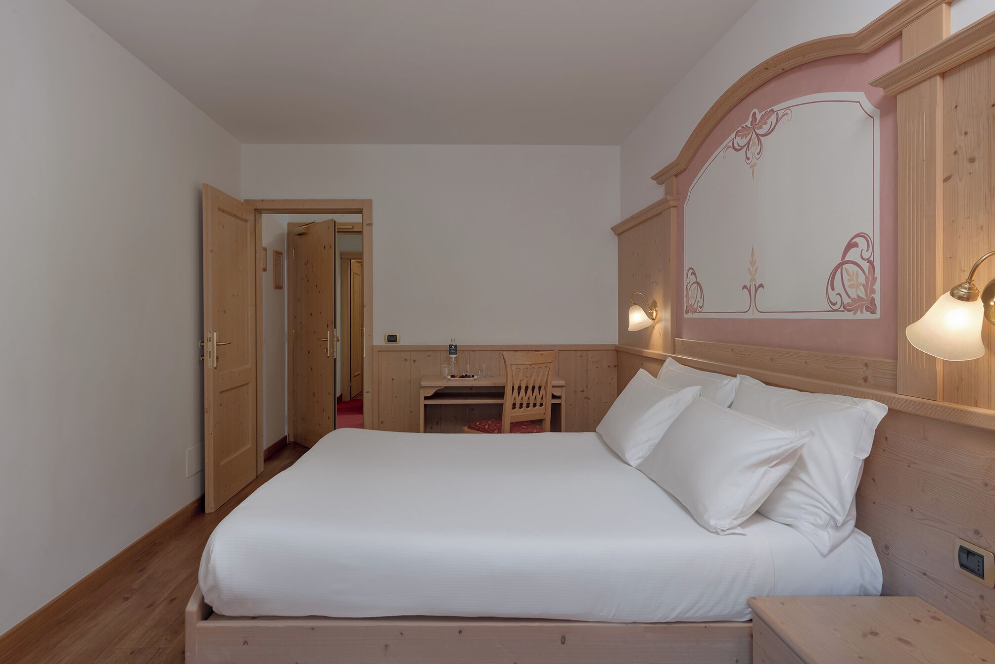 A bedroom with a large bed, wooden headboard, bedside lamps, and a small table with a chair.
