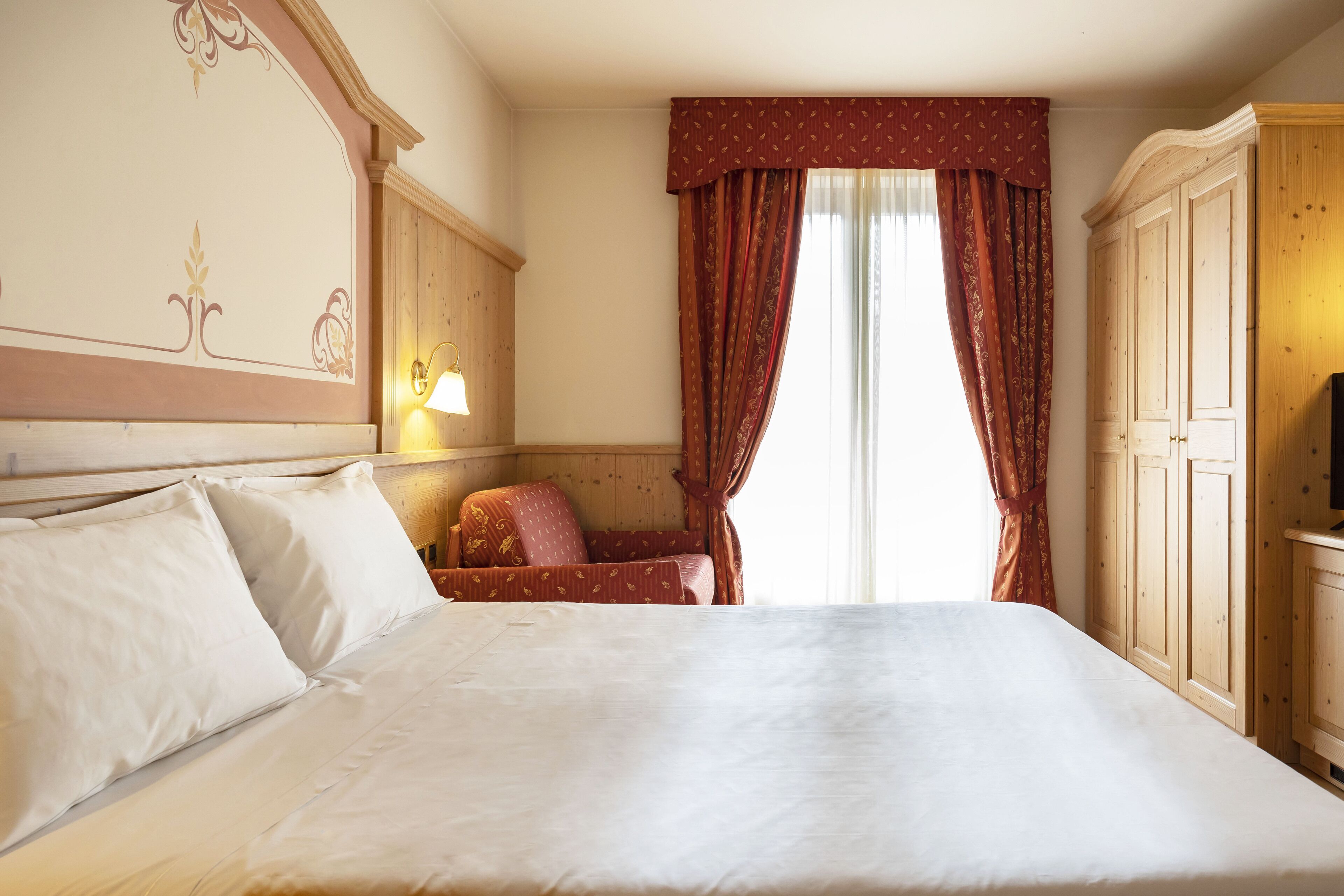 A hotel room with a large bed, a wooden wardrobe, a small armchair, and a window with curtains.