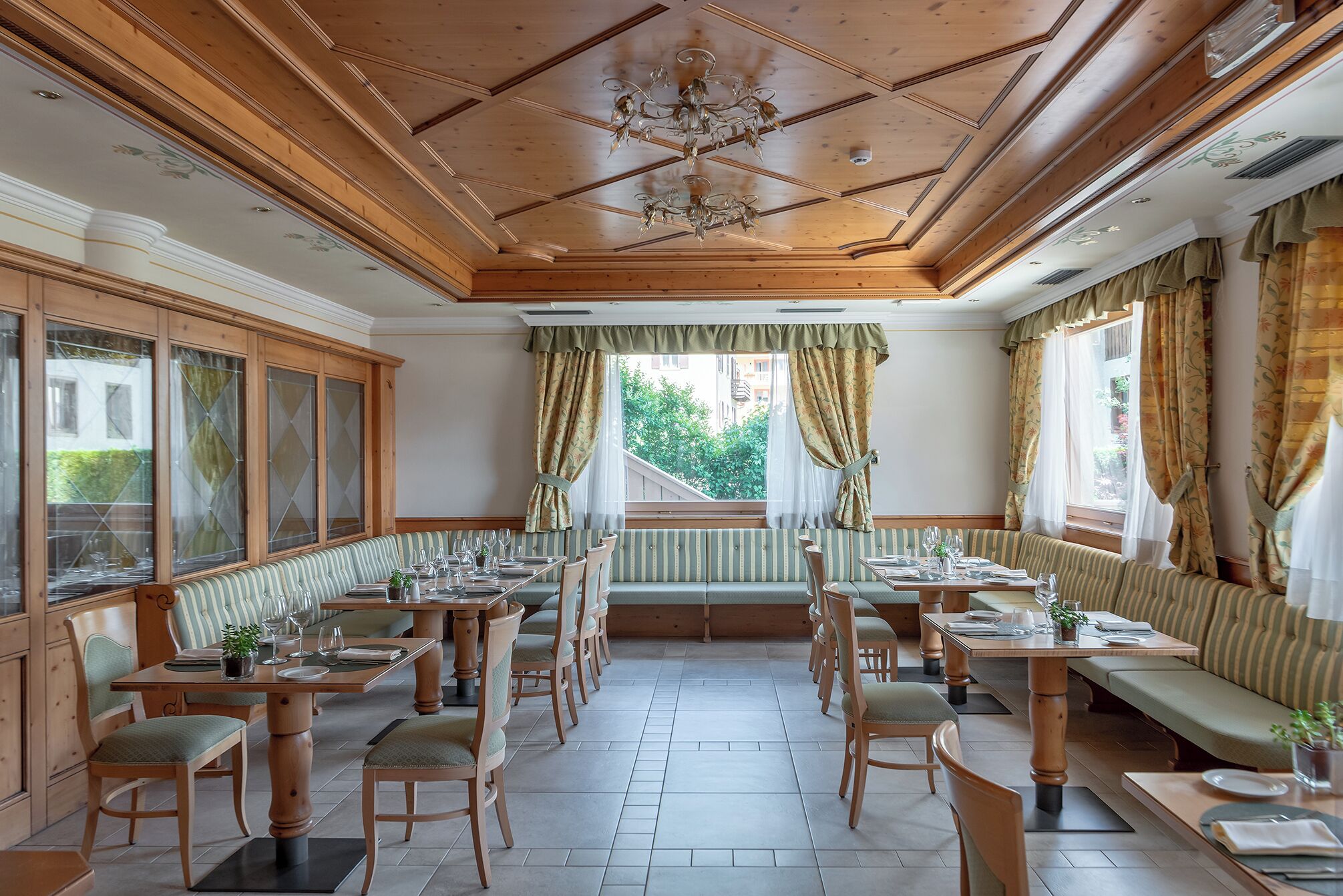 A dining area with wooden tables and chairs, a chandelier, and a view of greenery outside.