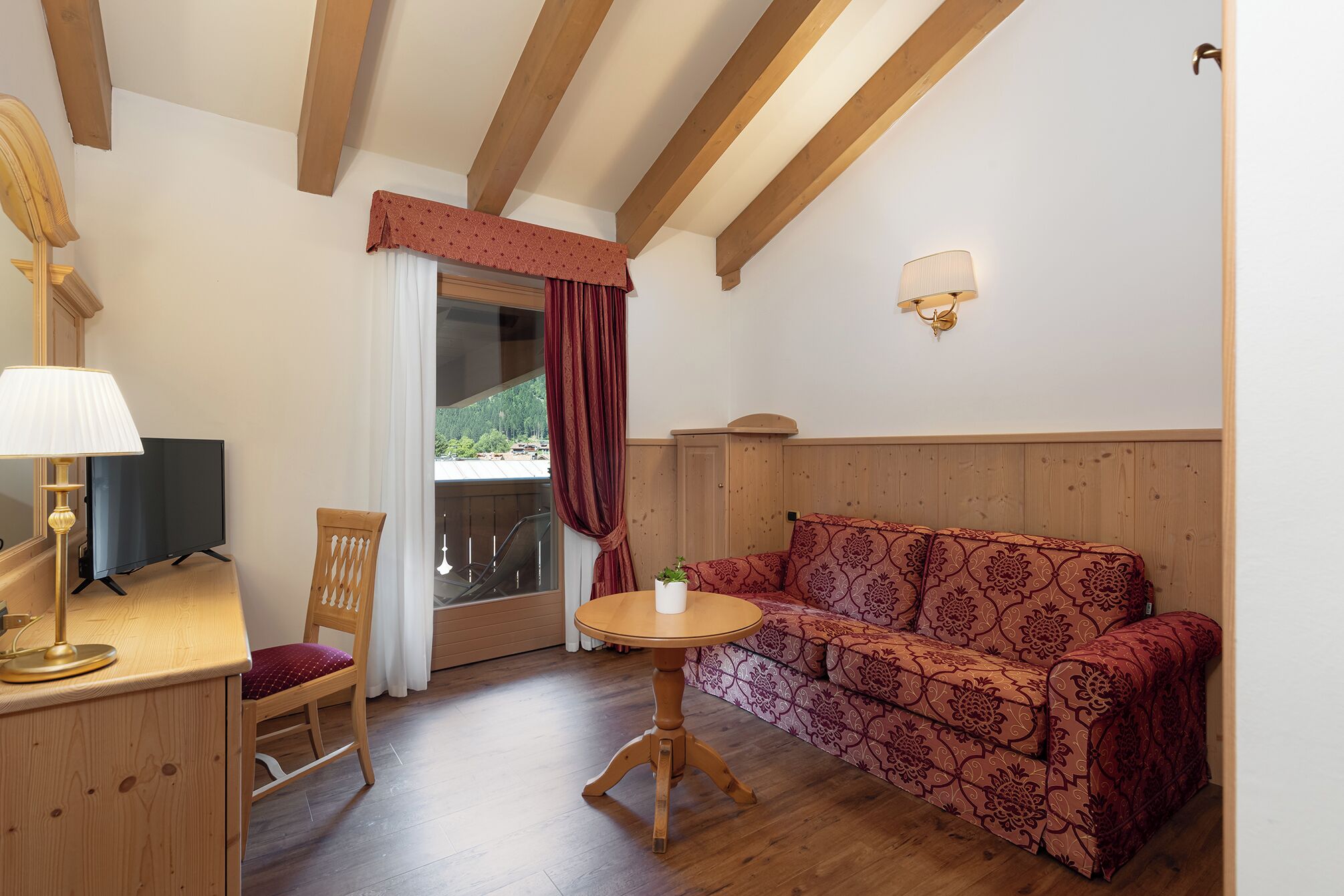 A room with a wooden ceiling, a sofa, a small table, a chair, a lamp, a TV, and a window with curtains.