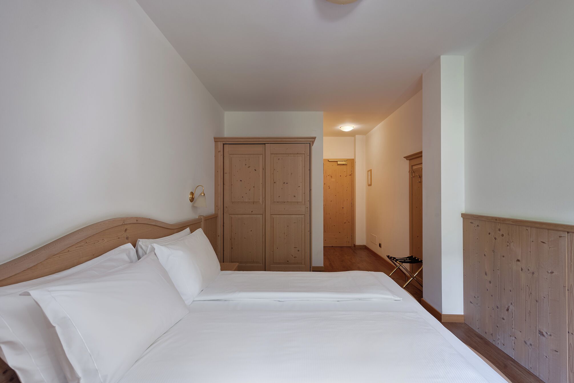 A bedroom with a wooden headboard, a bed with white linens, a wooden wardrobe, and a door leading to another room.