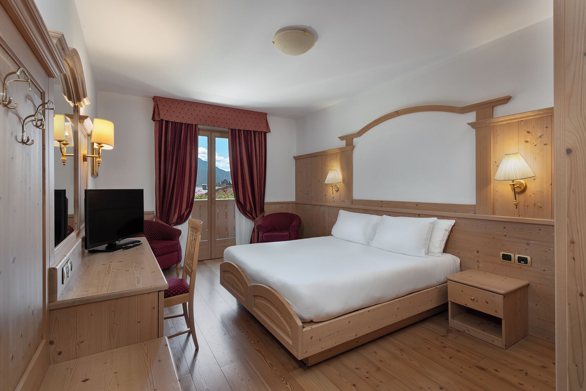 A hotel room with a wooden bed, a desk with a TV, a chair, and a view of mountains through the window.