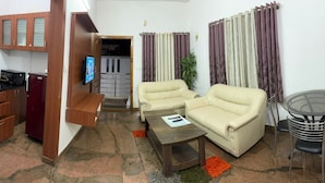 A compact living space with a kitchenette, two beige sofas, a coffee table, and a small dining table with chairs. - Nandu Hospitality – Aashiyana (Bengaluru)