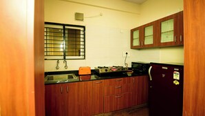 A compact kitchen with wooden cabinets, a sink, a stove, and a refrigerator. - Nandu Hospitality – Aashiyana (Bengaluru)