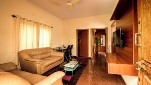 A living room with a sofa, a coffee table, and a TV. There is a wooden cabinet and a door leading to another room. - Nandu Hospitality – Aashiyana (Bengaluru)