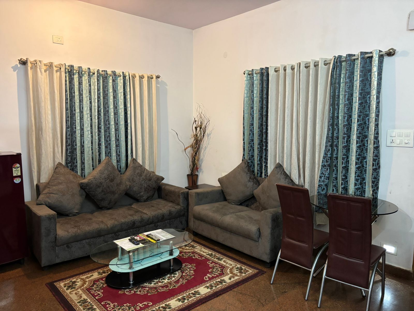 A living room with two sofas, a glass coffee table, a dining table with chairs, and patterned curtains.