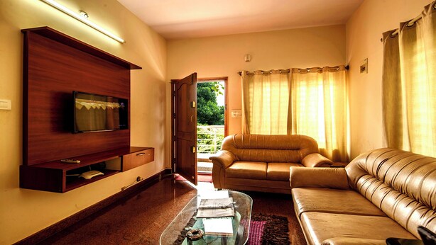 A living room with a TV, leather sofas, a glass coffee table, and a view of greenery outside. - Nandu Hospitality – Aashiyana (Bengaluru)