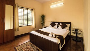 A hotel room with a large bed, a bedside table with a phone, a mirror, a window with curtains, and a small plant. - Nandu Hospitality – Aashiyana (Bengaluru)