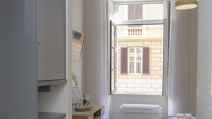 A compact bedroom with a bed, a desk with a chair, a wardrobe, and a window with a view of a building.
