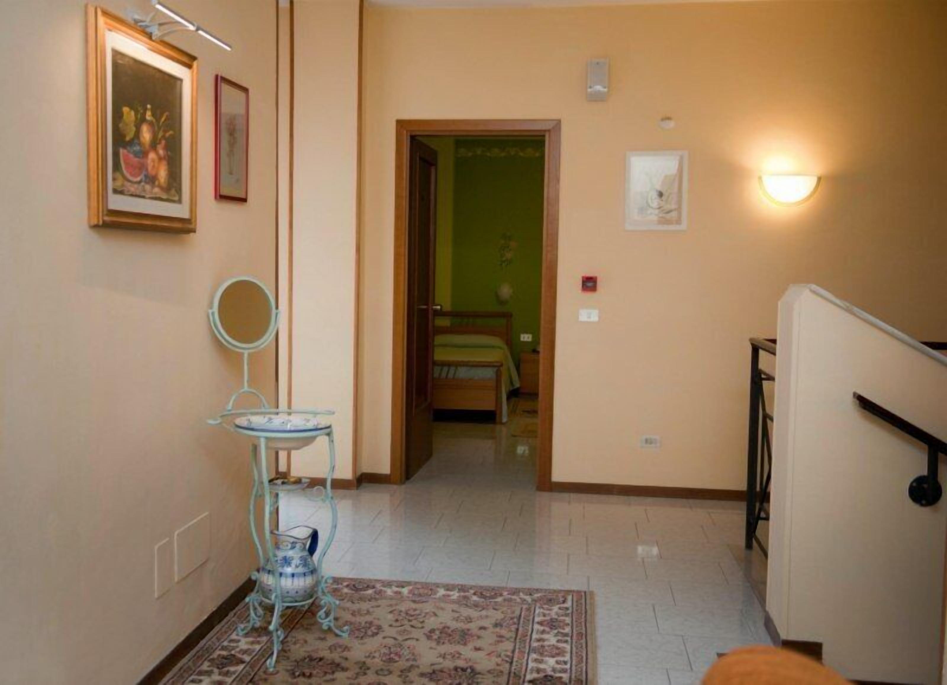 A hallway with a bed in the background, a small table with a vase, and a mirror on the wall.