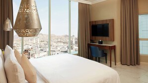 Ein Hotelzimmer mit einem großen Bett, einem Schreibtisch mit Stuhl, einem Fernseher und Blick auf eine Stadtkulisse durch große Fenster.