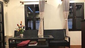 Una habitación con un sofá de mimbre negro, una mesa de centro de vidrio y una mesita con un jarrón de flores. Hay dos ventanas con cortinas y una puerta con manija.