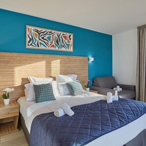 A hotel room with a large bed, a headboard, a sofa, a wall art piece, and a nightstand with a plant. - St Jean Luz Hotel Erromardie Suite 4p (Saint-Jean-de-Luz)