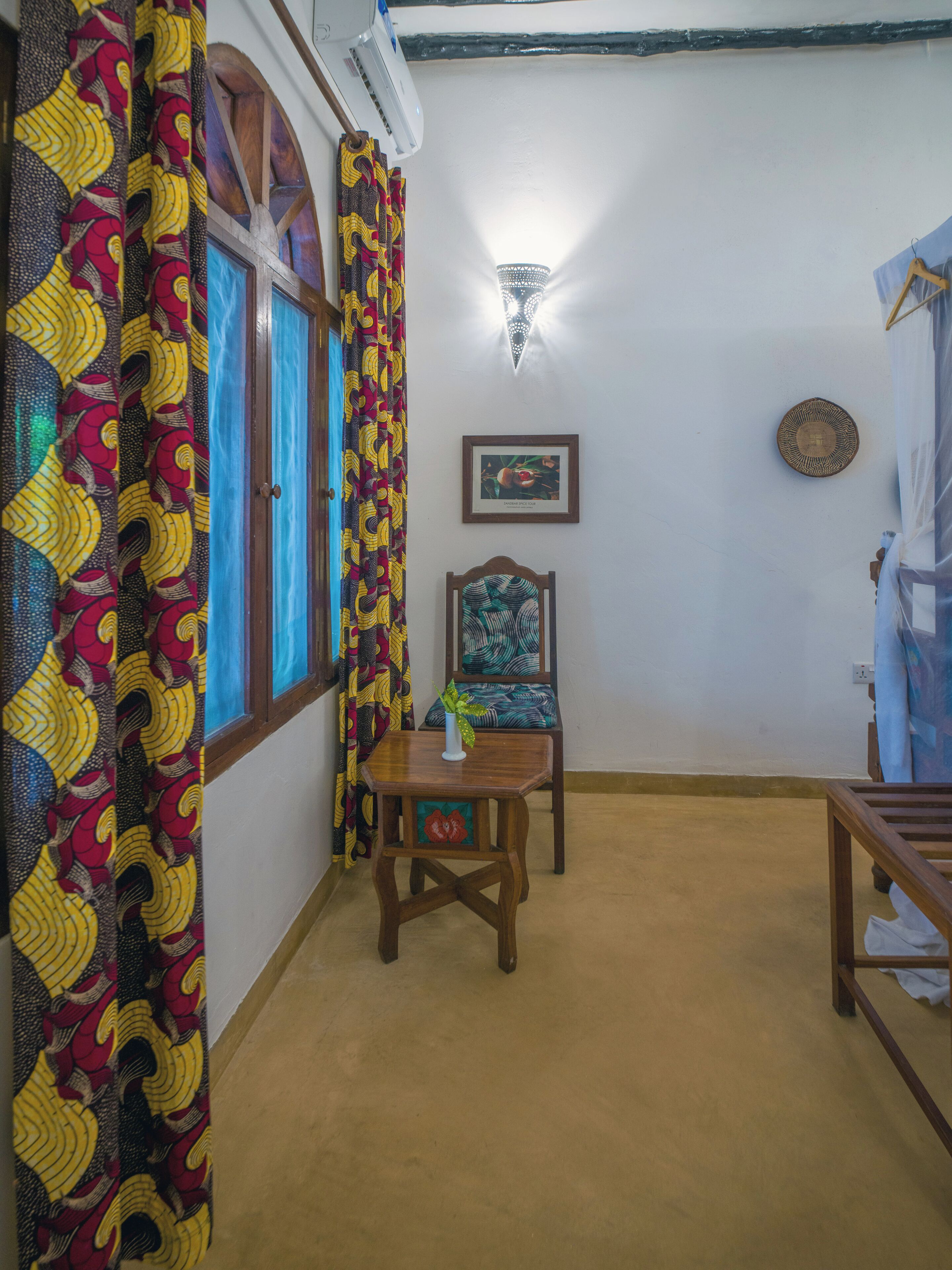 A room with a window, a wall-mounted light, a framed picture, and a small table with a vase.