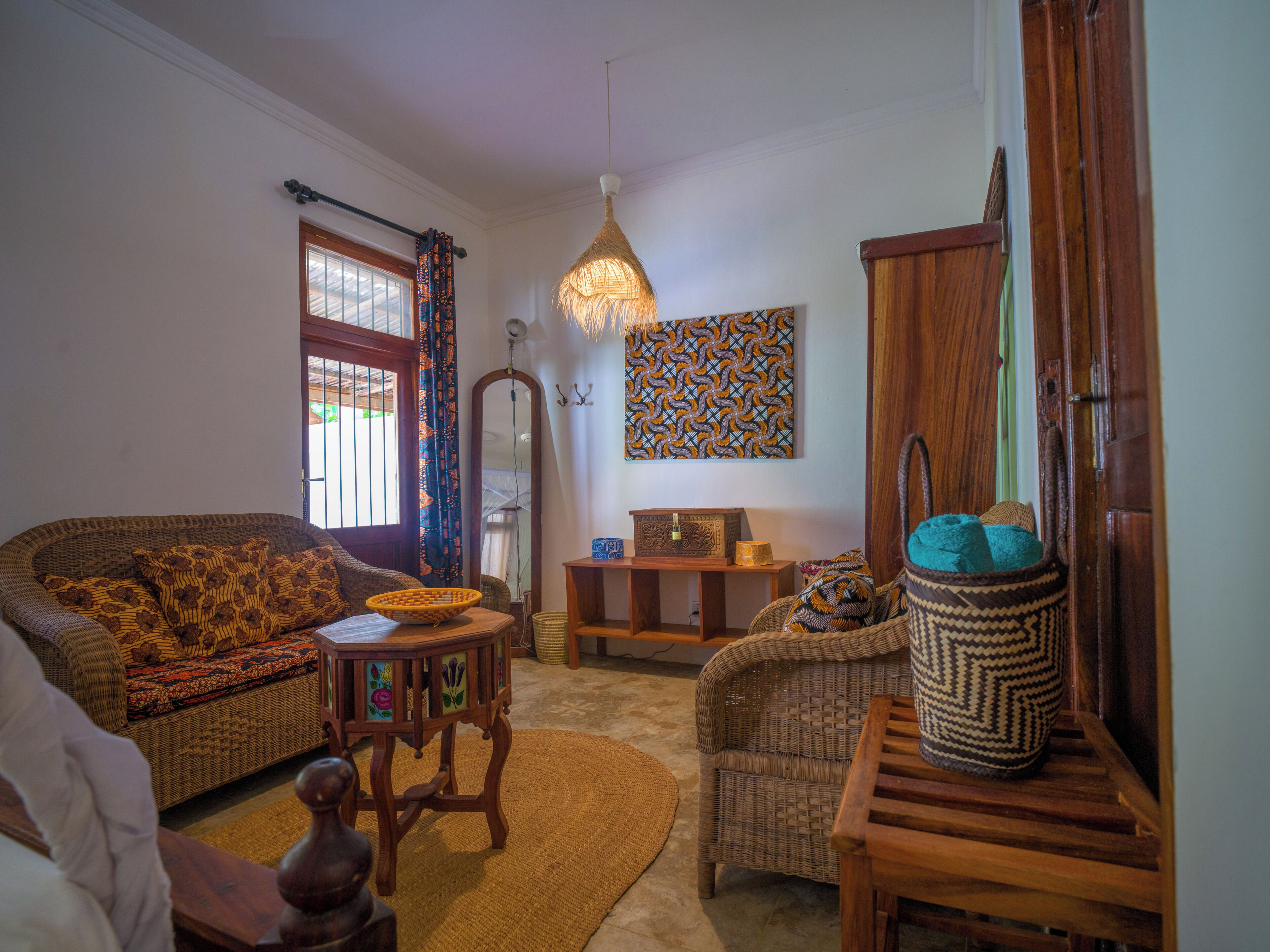 A living room with a sofa, a small table, a wicker chair, a hanging light fixture, and a patterned wall hanging.
