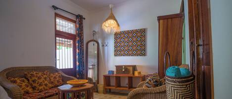 A living room with a sofa, a small table, a wicker chair, a hanging light fixture, and a patterned wall hanging.