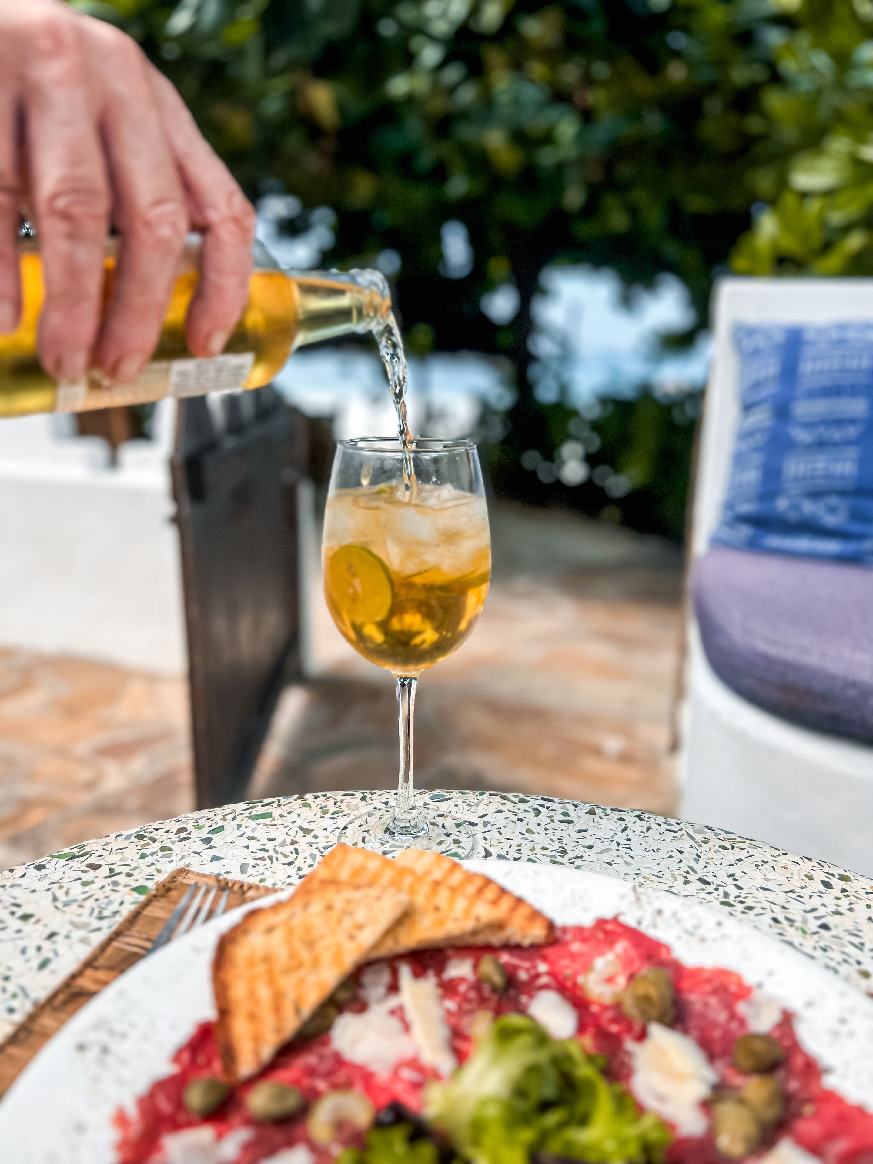 A glass of white wine being poured into a glass with ice and a slice of lime. A plate of food with various toppings is in front of the glass.