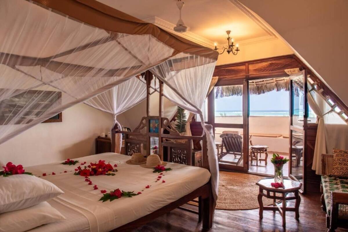 A bedroom with a bed, a nightstand, a chair, a table with a vase, and a view of the beach.