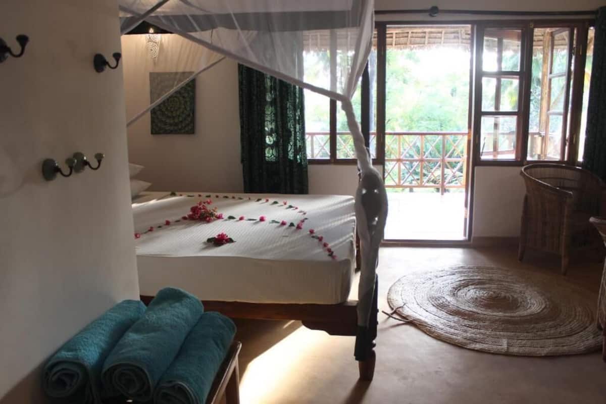 A room with a bed, a mosquito net, a chair, and a view of greenery through the window.