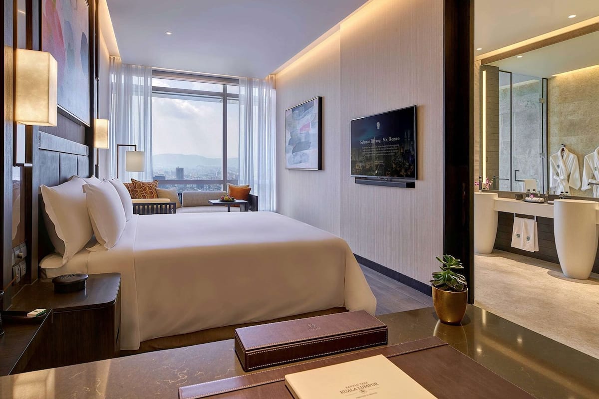 Habitación de hotel con una cama grande, mesitas de noche, un escritorio, una silla, un ventanal con vistas a la ciudad, un televisor montado en la pared y un baño con espejo y toallero.