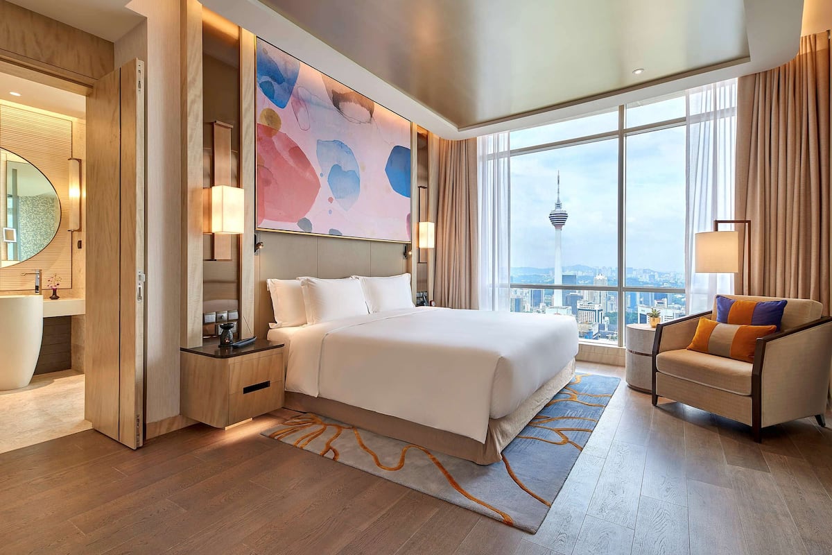 Habitación de hotel moderna con una cama grande, una zona de estar con una silla, vistas a la ciudad y un cuadro abstracto de grandes dimensiones en la pared.