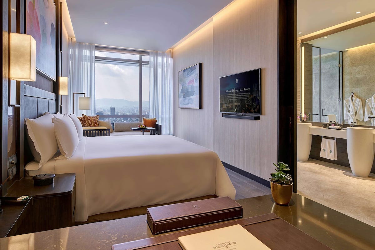 Una habitación de hotel moderna con una cama grande, un escritorio, una tocador con espejo y vistas de la ciudad a través de amplias ventanas.