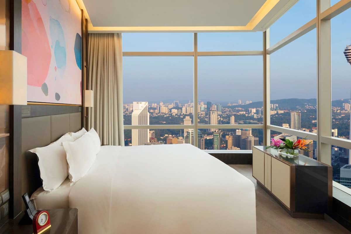 Habitación de hotel con una cama grande, una mesita de noche con un reloj, una ventana con vistas a la ciudad y un elemento decorativo en la pared.