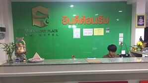 Een receptiebalie met een groene wand waarop de naam 'Thanakorn Place & Hotel' in zowel Thai als Engels staat vermeld. Op de wand hangen diverse borden en mededelingen, een kleine Boeddhabeeld en een plant aan de linkerzijde.