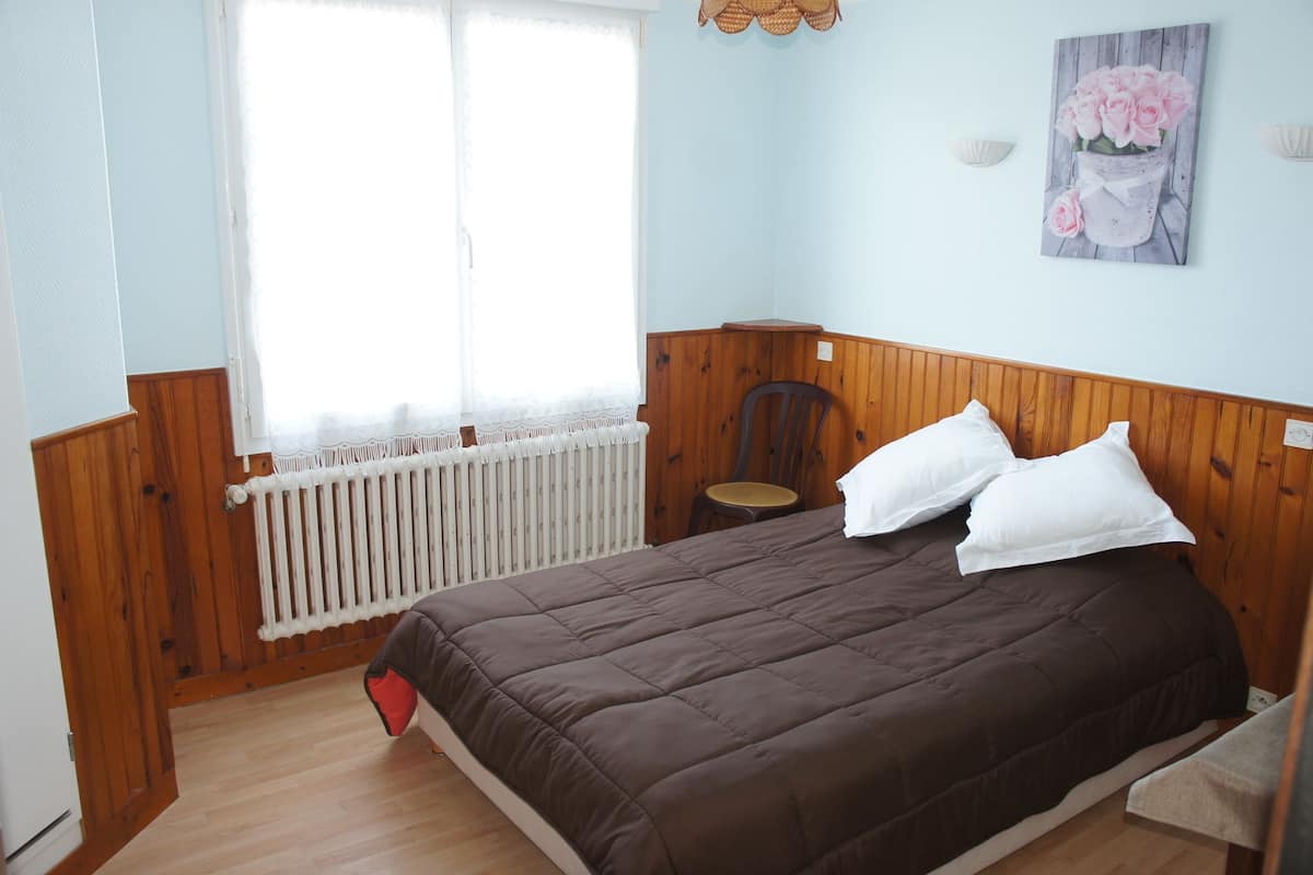 A bedroom with a wooden headboard, a brown bedspread, a white radiator, and a window with sheer curtains.