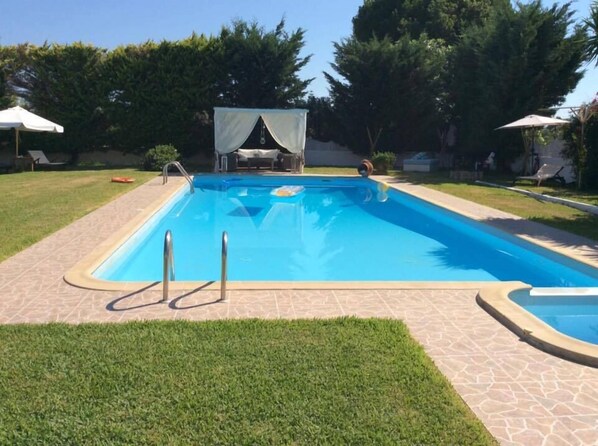 Une piscine rectangulaire, bordée d’une terrasse carrelée et entourée de pelouse et d’arbres.