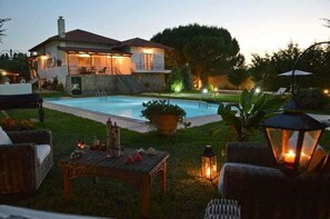 Una casa de dos pisos con piscina, mobiliario exterior y un jardín bien iluminado al anochecer.