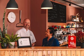 Uma área de recepção de hotel com balcão de madeira, um relógio, um menu em lousa e um certificado emoldurado.