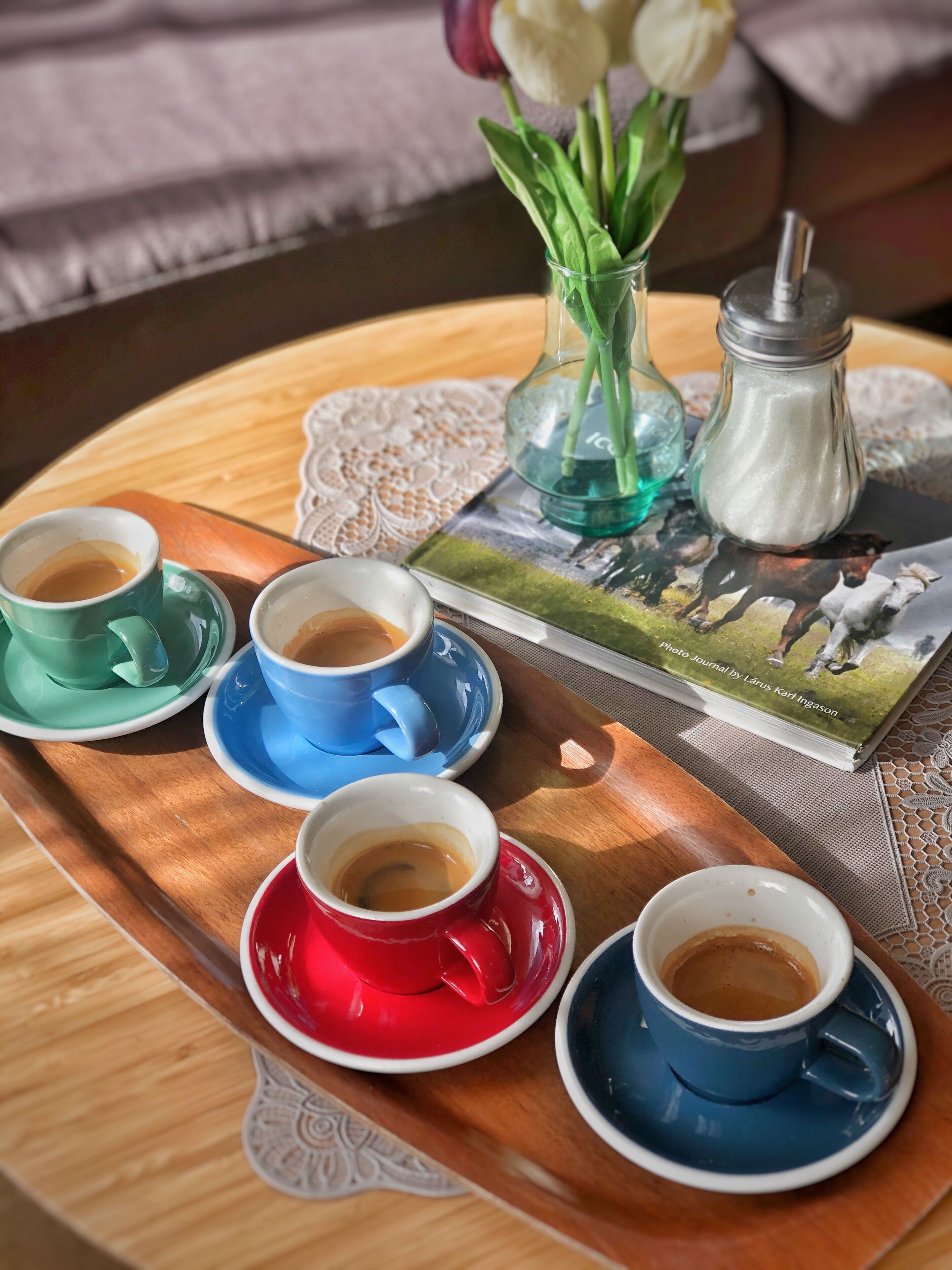 A tray with four cups of coffee, a salt shaker, a vase with flowers, and a photo journal on a wooden table.