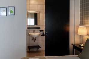 Quarto de hotel com uma cama, uma mesinha de cabeceira com abajur, uma porta preta, um banheiro com pia e espelho, e duas imagens emolduradas na parede.