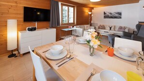 Ein gemütliches Blockhaus-Interieur mit Holzwänden, einer Essecke mit weißem Geschirr, einer Couch und einem an der Wand befestigten Fernseher.